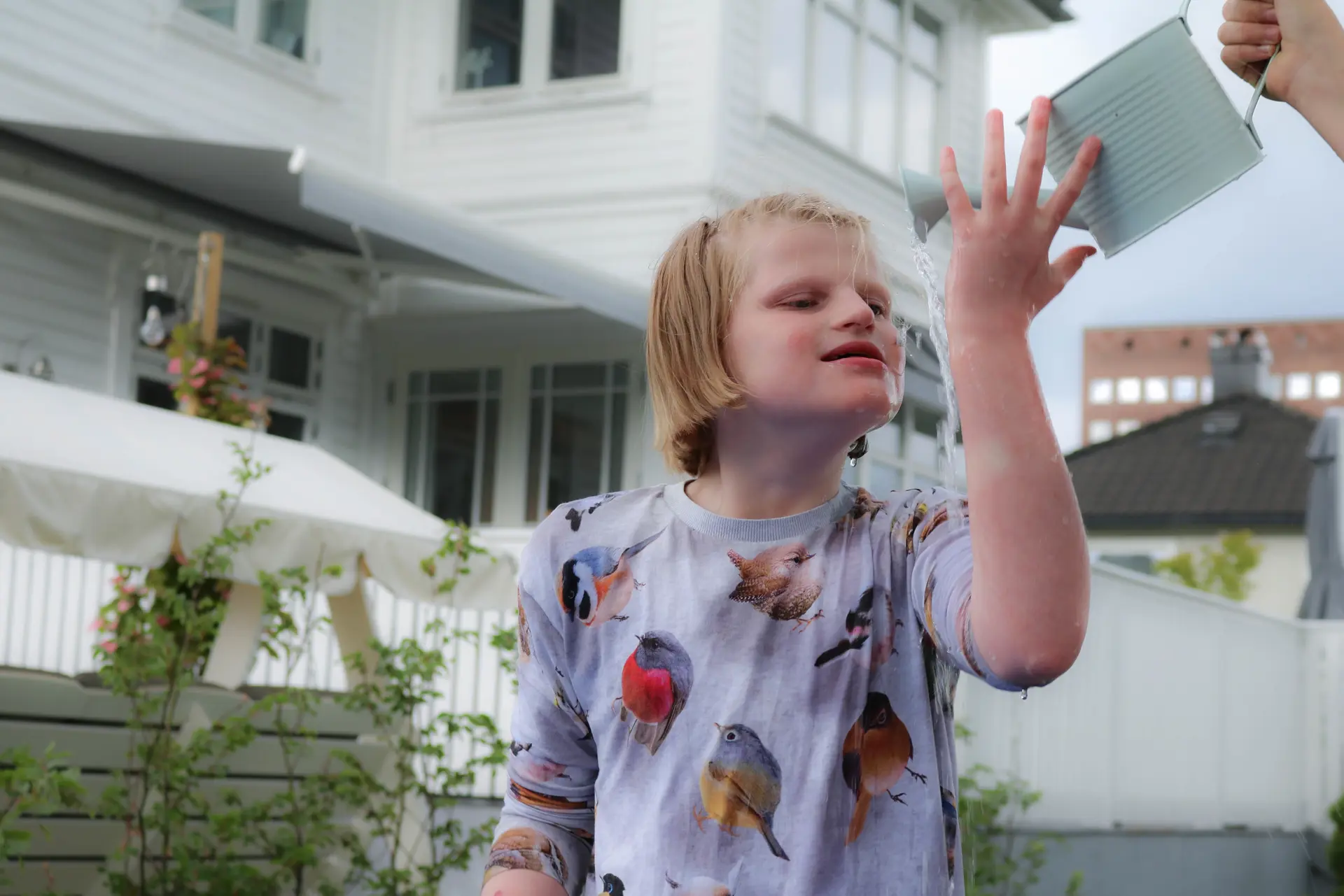 Jente står foran hvit villa, hun holder venstrehånden opp mot en vannkanne. Det renner vann nedover armen hennes.
