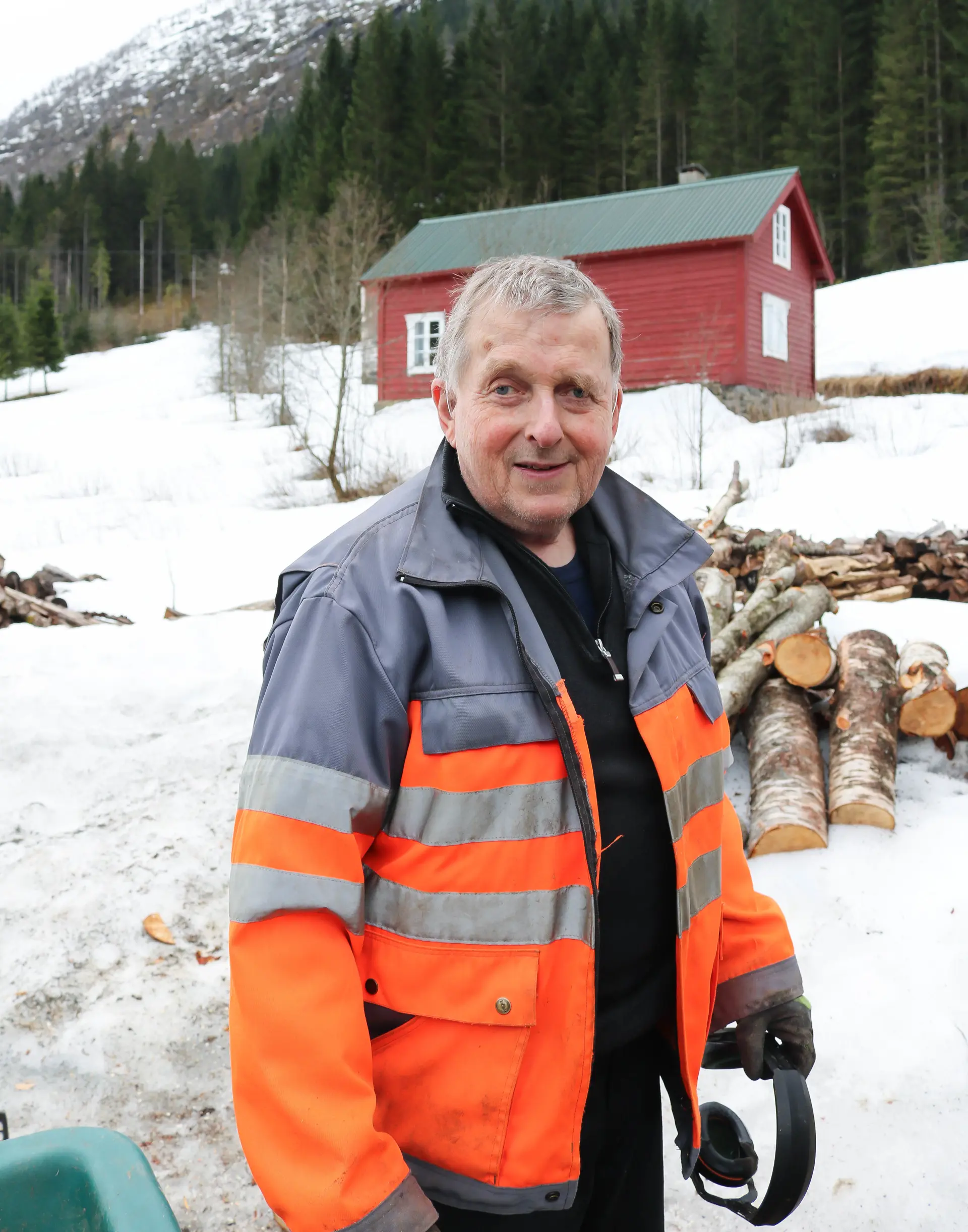 En mann iført en oransje jakke og stående foran noen vedstabler i snøen